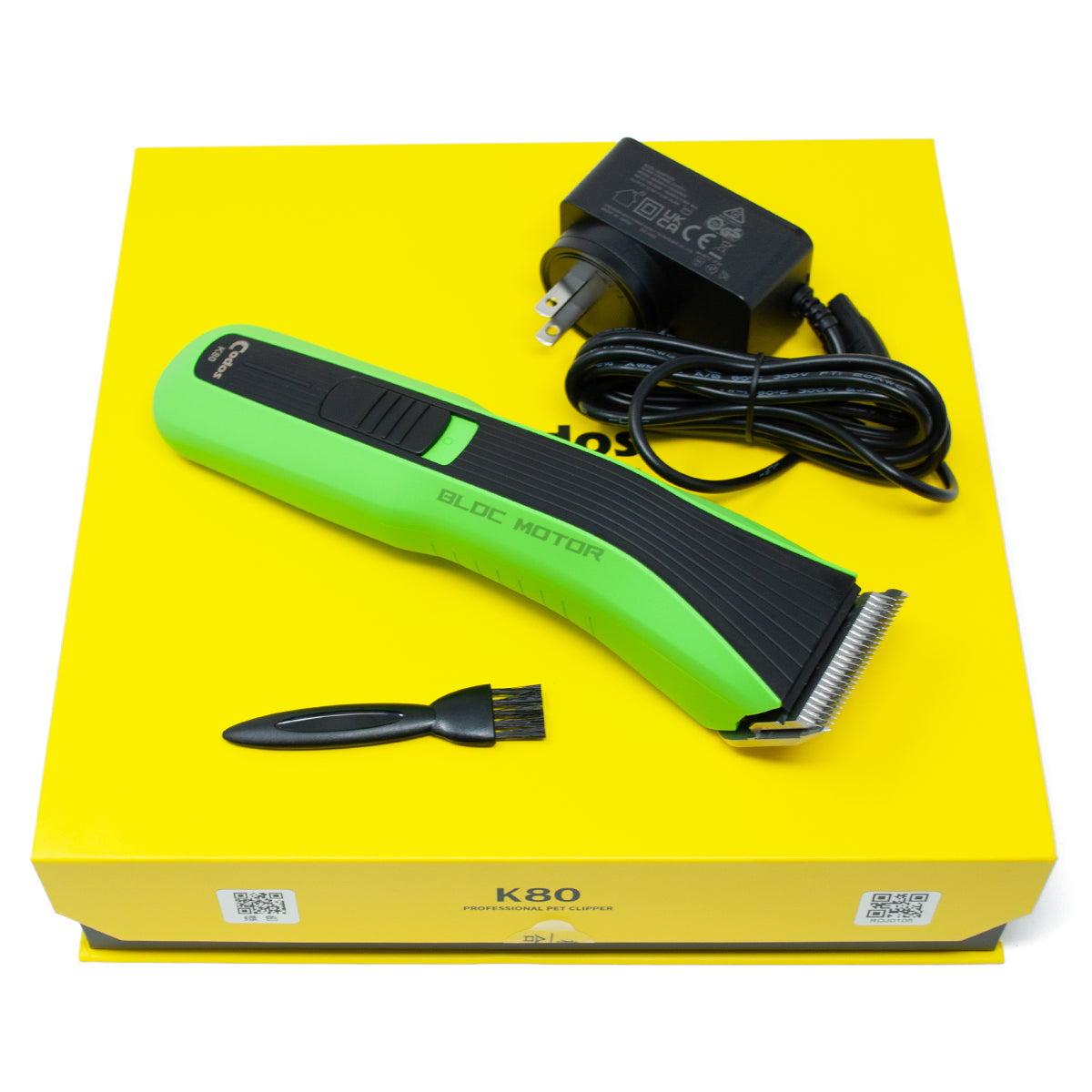 A green and black cordless electric hair clipper set displayed on a bright yellow box, with the matching black power adapter and a small black cleaning brush included.
