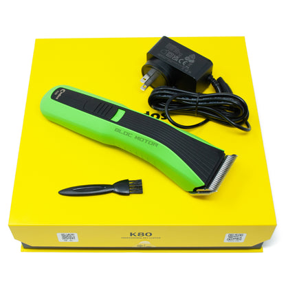 A green and black cordless electric hair clipper set displayed on a bright yellow box, with the matching black power adapter and a small black cleaning brush included.